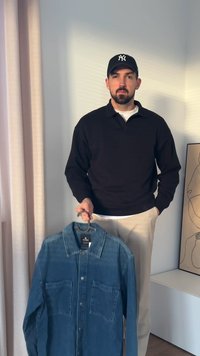 Denim-paita, jossa on nappikiinnitys, kaksi etutaskua ja klassinen kaulus, keskisinisenä. Pitäjänä on henkilö mustassa neuleessa.