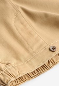 Tissu beige clair, présentant une texture douce, un ourlet froncé et un accent de bouton marron. Coutures visibles autour des bords.