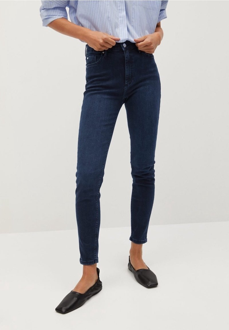 Personne ajustant un jean skinny bleu foncé associé à des ballerines pointues noires et une chemise rayée bleu clair.