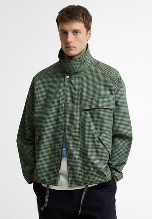 Noor mees, kes kannab rohekat ja pruunikat ruudulist jopet kõrge kraega ja suruöökidega, käed taskutes, seisab valge tausta ees.