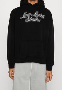 Sweat-shirt à capuche noir avec l'inscription "Low Lights Studios" brodée en lettres grises, doté d'une poche kangourou à l'avant et de poignets côtelés.