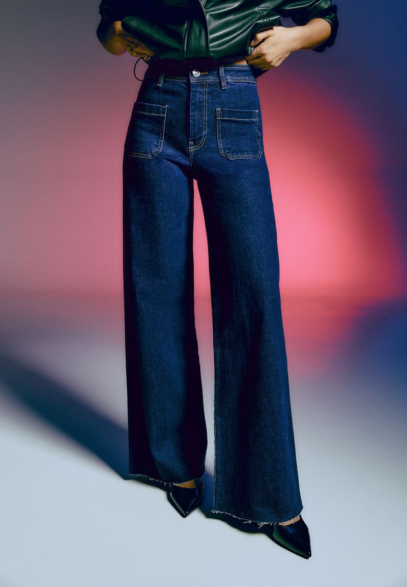 Jean en denim foncé à jambe large et taille haute, doté de quatre poches, de coutures visibles et d'accents de bord effiloché. Porté avec des talons aiguilles noirs.