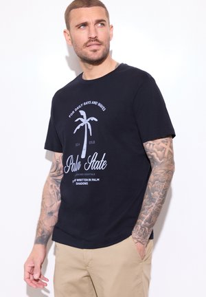 Man met getatoeëerde armen die een zwarte "Palm State" T-shirt en beige broek draagt, kijkt naar links tegen een witte achtergrond.