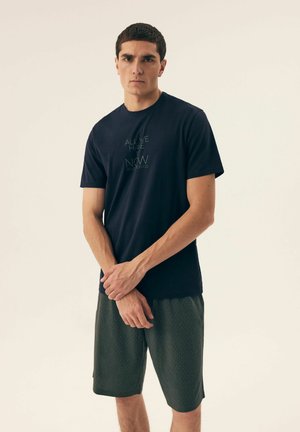 Dunkelblaues T-Shirt mit kurzen Ärmeln, versehen mit grünem Textdruck. Kombiniert mit gemusterten grünen Shorts hat das Modell einen neutralen Gesichtsausdruck.