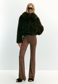 Donna che indossa un giubbotto di pelliccia sintetica marrone scuro, pantaloni marroni a zampa d'elefante con spacchi alla caviglia, tacchi aperti in pelliccia coordinata e grandi occhiali da sole scuri.