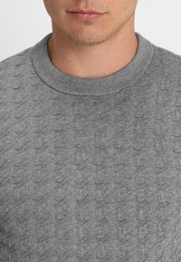 Il maglione grigio in maglia presenta un motivo testurizzato, un colletto rotondo e un orlo a costine. Il tessuto appare morbido e leggero.