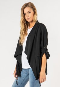 Zwarte lichte wikkeljas met gedrapeerde voorkant, lange mouwen, knoop manchetten en verstelbare strikdetails in de taille, gedragen over een witte blouse.