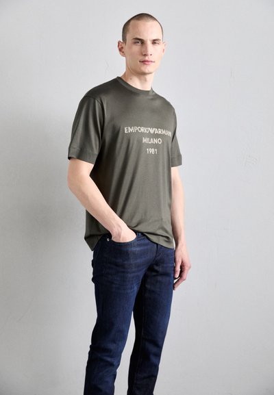 Tamsiai žalia medvilnės marškinėlių su apvalia iškirpte ir trumpomis rankovėmis, ant kurių yra balta užrašas "EMPORIO ARMANI MILANO 1981." Dėvimi su tamsiai mėlynomis džinsomis.