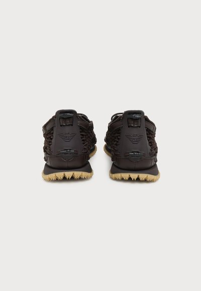 Paire de sandales tissées marron foncé avec semelles en caoutchouc beige, vues de dos sur un fond clair uni.
