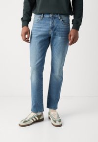 Lichtblauwe denim jeans met een rechte snit en subtiele vervaagde plekken, gecombineerd met grijsgroene sneakers met crèmekleurige veters en bruine accenten.