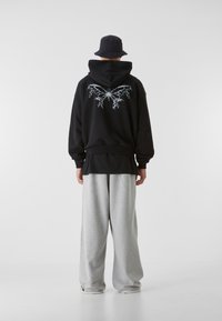 Personne portant un chapeau seau noir, un sweat à capuche noir avec un motif de papillon blanc dans le dos, et un pantalon de survêtement gris ample, debout devant un fond uni.