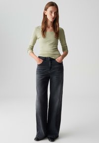 Lichtgroene blouse met lange mouwen en zwarte wijde jeans. De jeans hebben zijnaad en een gladde textuur, terwijl de blouse een licht getailleerde snit heeft.