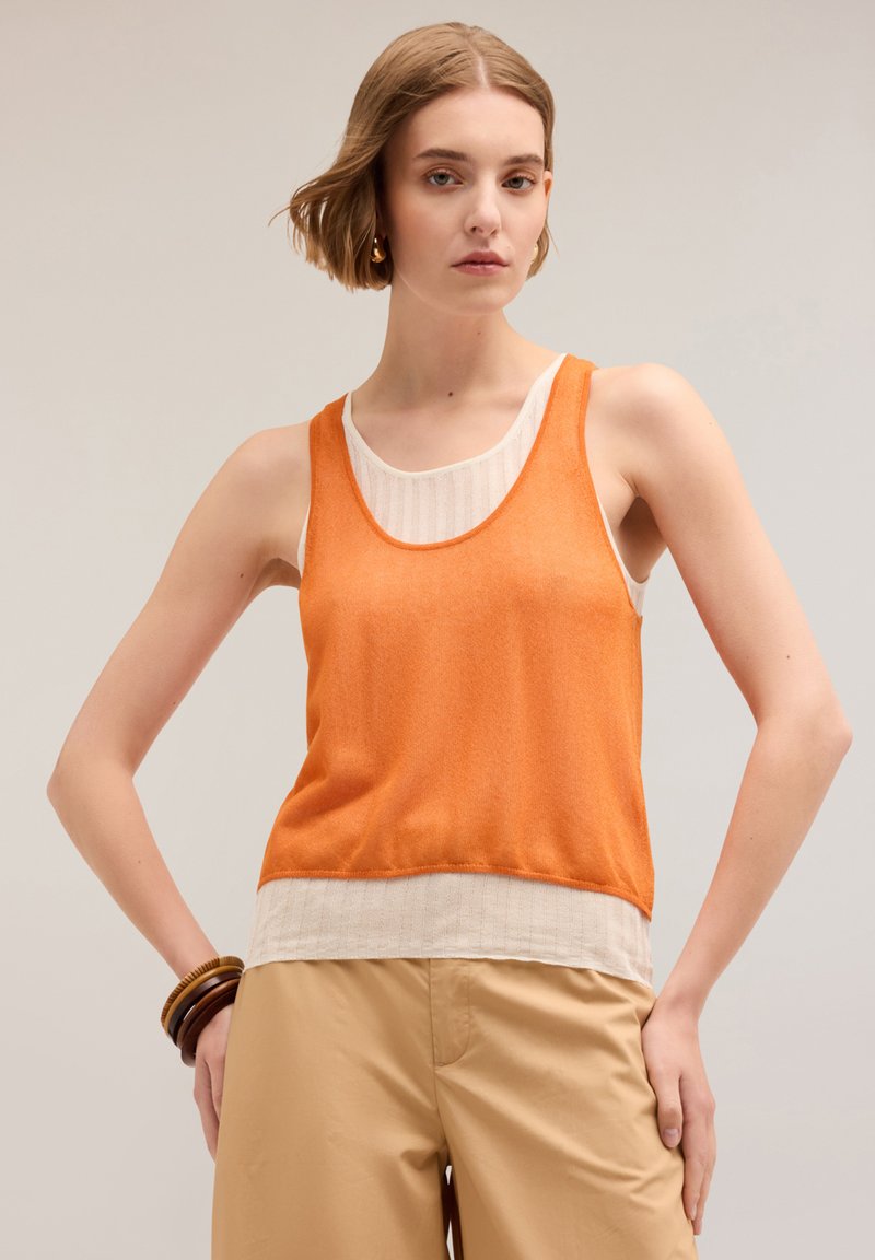 Top sin mangas de color naranja con un escote redondo sobre una capa blanca de canalé, combinado con pantalones beige. Textura suave, ajuste casual.