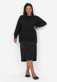 Zwarte, gestructureerde trui met sprankels, gecombineerd met een zwarte midi-rok met een split aan de voorkant. Zwarte schoenen met strikaccenten maken de look compleet.