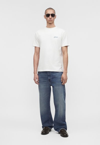 Άνδρας που φοράει λευκό κοντομάνικο μπλουζάκι, άνετο μπλε τζιν, μαύρα παπούτσια και σκοτεινά γυαλιά ηλίου, στέκεται μπροστά σε απλό φωτεινό φόντο.