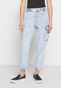 Osoba ubrana w jasnoniebieskie spodnie dresowe z zasłoniętymi kieszeniami, z wiązaniem w pasie i czarnymi butami na ślizg, stojąca na tle białym.