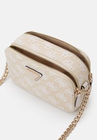Krémová crossbody taška s texturovanou látkou, dvojitými zipovými přihrádkami, zlatými kovovými akcenty a řetízkovým popruhem. Obsahuje detail s logem.