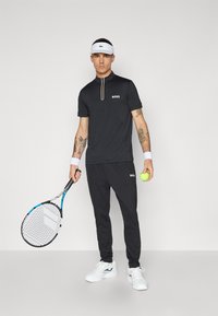Svart kortärmad polo med dragkedja och vit "BOSS"-logotyp, matchad med svarta joggingsbyxor, vita tennisskor, och hållande en tennisracket och boll.