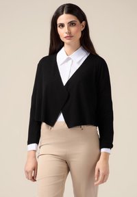Svart kortärmad cardigan i texturerat tyg med omslagdesign och långa ärmar. Bärs över en vit skjorta med krage och beige byxor.