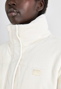 Gilet en velours côtelé blanc avec col montant, arborant un petit patch logo CK. Le tissu présente un motif texturé et une apparence douce.