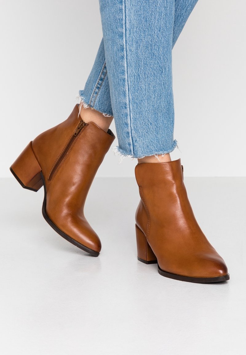 Bottes en cuir marron à talons blocs portées avec un jean bleu clair à ourlet effiloché sur un sol blanc.