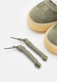 Green suede shoes with a textured upper, beige rubber soles, and round laces lying alongside them. Laces are tightly wrapped and knotted.