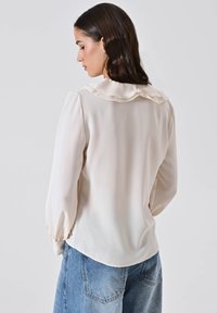 Donna con capelli scuri che indossa una blusa crema con colletto a volant e jeans in denim blu, vista da dietro su uno sfondo chiaro e neutro.