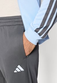 Αθλητικό μπουφάν ανοιχτό μπλε με γκρι φόρμα. Το γκρι παντελόνι φέρει λευκό λογότυπο Adidas· το μπουφάν έχει σκούρες γκρι ρίγες κατά μήκος των μανικιών.