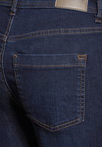 Mörkblå denimjeans med en bakficka, som har ljusbrunt sömnad och en läderlapp vid midjan. Slät textur.