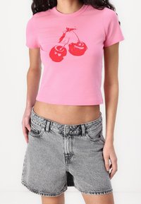 Kvinna klädd i en rosa kortärmad t-shirt med röd körsbärsgrafik, tillsammans med högmidjade, blekta grå denimshorts.