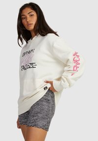 Cream hoodie with black and pink text graphics, kangaroo pocket, ribbed cuffs; paired with striped black and white shorts.