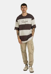 Kastige braun-beige gestreifte T-Shirts mit gesticktem Logo, kombiniert mit beigen Cargo-Hosen mit Seitentaschen und elastischen Bündchen. Weiße Sneaker.