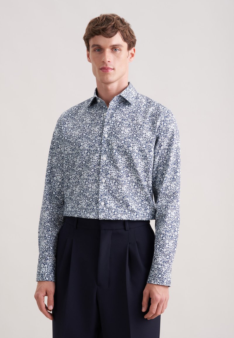 Jeune homme portant une chemise à manches longues bleue et blanche à fleurs, boutonnée et rentrée dans un pantalon noir taille haute, se tenant devant un fond uni.