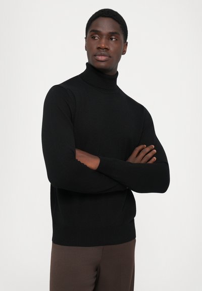 Pulover negru cu guler înalt, din material tricotat, design ajustat cu mâneci lungi și manșete cu nervuri. Modelul pozează cu brațele încrucișate, pe un fundal neutru.