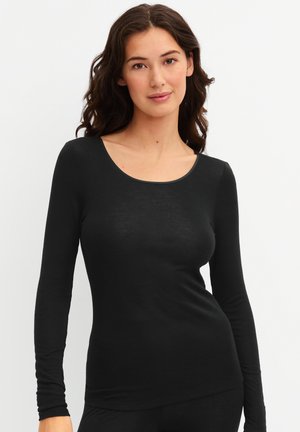 Femme aux longs cheveux bouclés portant un haut noir moulant à manches longues, debout devant un fond clair uni.