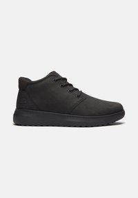Chaussure noire montante à lacets avec semelle texturée et col matelassé, logo embossé sur le côté près du talon, sur un fond clair uni.