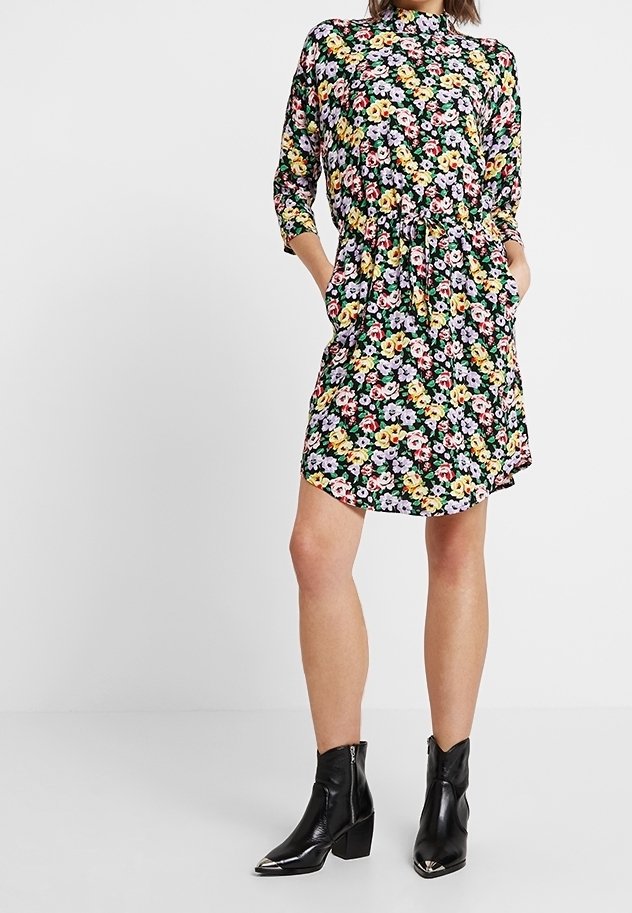 Femme portant une robe fleurie à manches longues et longueur genou, les mains dans les poches, avec des bottines noires à bouts métalliques.