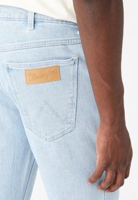 Světle modré džíny z denimu s koženou nášivkou "Wrangler" na zadní kapse, která má výrazný prošívaný vzor ve tvaru 'W'.