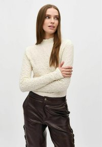 Geverfde crèmekleurige lange mouwtop met een hoge hals en een aansluitende silhoët, gecombineerd met donkerbruine, glanzende, gemusterde leren broek.
