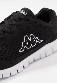 Zapato deportivo negro con parte superior de malla, cierre con cordones y logo Kappa blanco prominente. Suela de goma blanca con patrón de agarre texturizado.