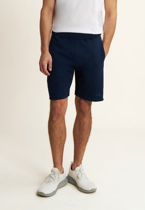Pantaloni sportivi blu navy con vita elastica, realizzati in un materiale morbido, abbinati a sneakers bianche con suola grigia chiara.