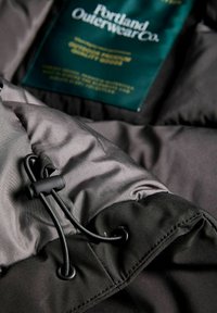 Czarna kurtka puchowa z gładką, błyszczącą powierzchnią. Posiada regulowane sznurki i metkę z napisem "Portland Outerwear Co."