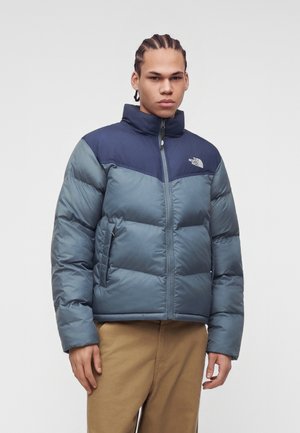 Joven con el pelo trenzado que lleva una cazadora acolchada de North Face azul y azul marino y pantalones beige, de pie frente a un fondo liso y claro.
