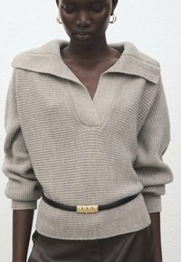 Maglione grigio chiaro lavorato a maglia con collo ampio e scollo a V profondo, caratterizzato da un motivo testurizzato e una cintura nera con un accento dorato in vita.