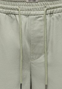 Jasnozielone spodnie z elastycznym stanem, ze sznurkami do ściągania i widocznymi szwami, pokazujące przednią część pasa i część zamka.