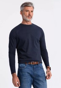 Námořnické tričko s dlouhým rukávem vyrobené ze 100% bavlny, kulatý výstřih, přiléhavý střih. Postaveno v kombinaci s světle modrými džíny a hnědým opaskem, na jednobarevném pozadí.