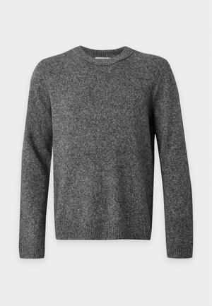 Ung mand iført en mørkegrå strikket sweater, lyseblå jeans med vide ben og sorte sko, står mod en ensfarvet lys baggrund.
