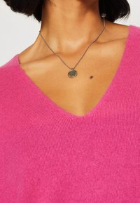 Suéter rosa de cuello en V con textura acanalada, llevado con un collar de pendant circular en plata y oscuro. El tono de piel es visible por debajo del escote.