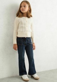 Cardigan bianco lavorato a maglia con un motivo testurizzato, abbinato a jeans a zampa d'elefante blu scuro con tasche frontali e cuciture a contrasto. Scarpe bianche.