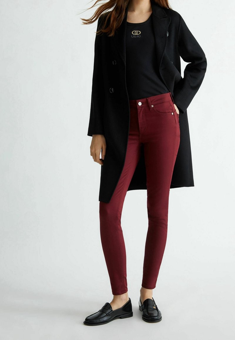 Femme portant un manteau noir, un top noir avec un logo doré, un pantalon slim bordeaux et des mocassins noirs, debout devant un fond clair uni.